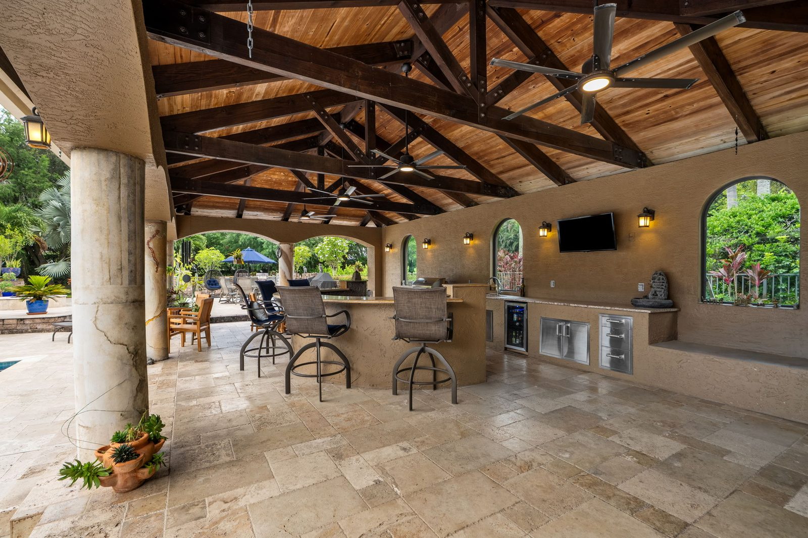 Covered outdoor living room under a wood-truss ceiling with summer kitchen and bar